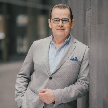 corma- Ein Mann mit Brille und grauem Blazer und blauem Hemd steht mit den Händen in den Taschen an einer Wand lehnend in einer städtischen Umgebung.
